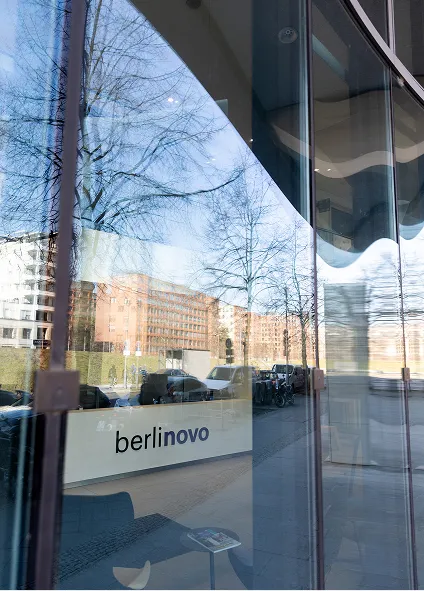 Foto von außen durch die Glasfront des Bürogebäudes in der
Linkstraße. Im Inneren ist der Empfangstresen mit der Aufschrift
„berlinovo“ zu erkennen.
