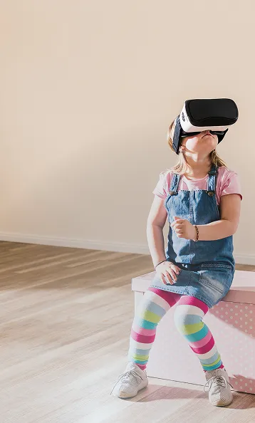 Hero Image Ein kleines Mädchen in Ringelstrumpfhose sitzt auf einem Hocker mit
einer VR-Brille auf dem Kopf und schaut gespannt nach oben.