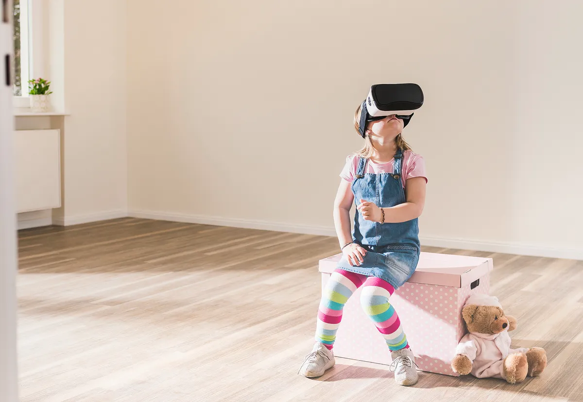 Hero Image Ein kleines Mädchen in Ringelstrumpfhose sitzt auf einem Hocker mit
einer VR-Brille auf dem Kopf und schaut gespannt nach oben.