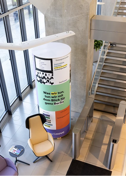 Blick hinunter in die Lobby des modernen Bürogebäudes in der
Linkstraße. Ein Loungesessel, eine Rolltreppe und eine Litfaßsäule
sind zu sehen.