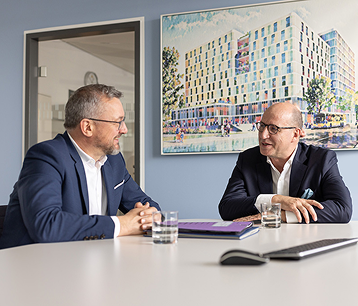 Geschäftsführer Alf Aleithe und Bereichsleiter Wolfgang Falk sind an
einem Tisch sitzend ins Gespräch vertieft. Hinter ihnen an der Wand
hängt das Bild eines großen modernen Neubaus.