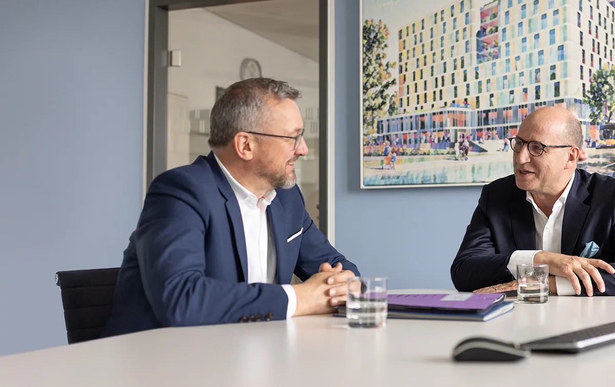 Geschäftsführer Alf Aleithe und Bereichsleiter Wolfgang Falk sind an
einem Tisch sitzend ins Gespräch vertieft. Hinter ihnen an der Wand
hängt das Bild eines großen modernen Neubaus.