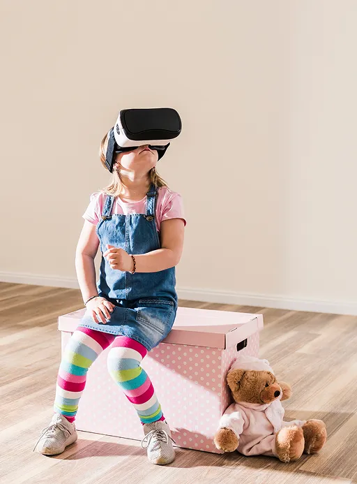 Ein kleines Mädchen in Ringelstrumpfhose sitzt auf einem Hocker mit einer VR-
Brille auf dem Kopf und schaut gespannt nach oben.