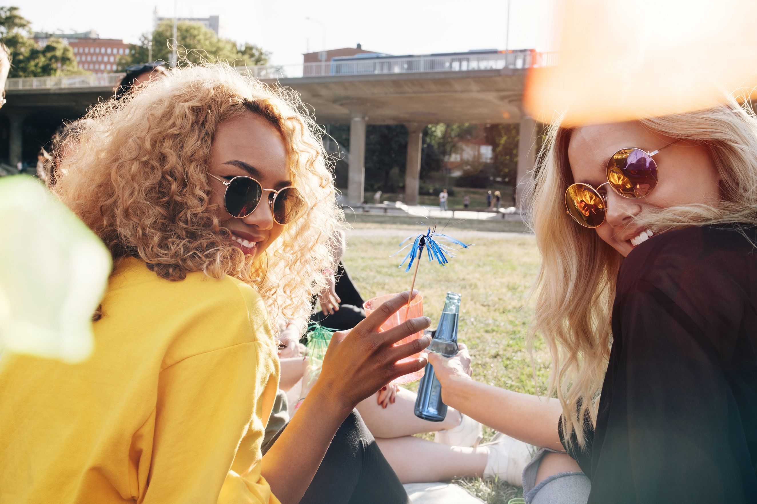 Hero Image Zwei junge Frauen mit Sonnenbrillen sitzen im Freien mitten in Berlin, schauen
über die Schulter zur Kamera und lachen fröhlich.