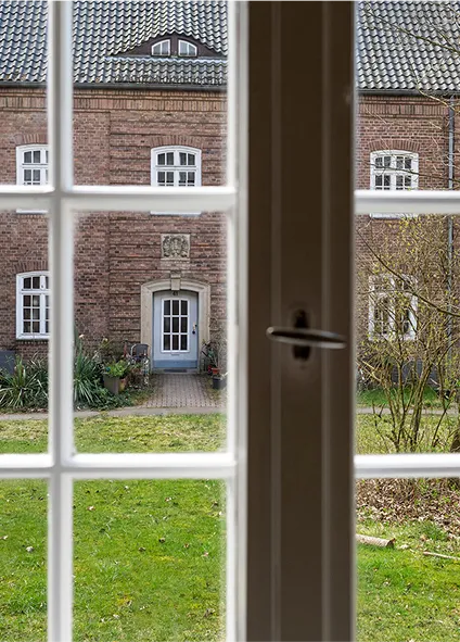 Blick aus einem weißen Sprossenfenster hinaus in den Garten hinüber
zu einem Klinkerwohnhaus der 30er-Jahre.