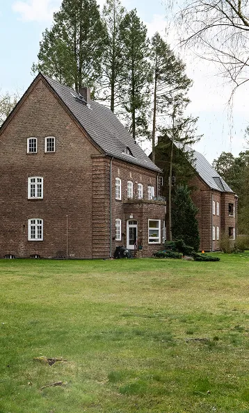 Hero Image Gartenansicht auf doppelstöckige rote Klinkerbauten umrahmt von
Bäumen.